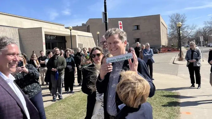 A group of people gathered outdoors, with James Ehnes holding a "James Ennes Way" street sign on a sunny day.