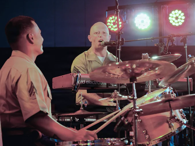Marine musicians perform on stage during a live concert, with a drummer and keyboard player singing into microphones under colorful stage lights.