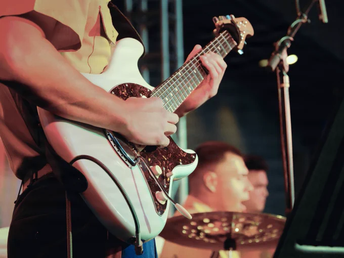 A Marine musician plays an electric guitar on stage during a live performance, with fellow band members and stage lighting visible in the background.