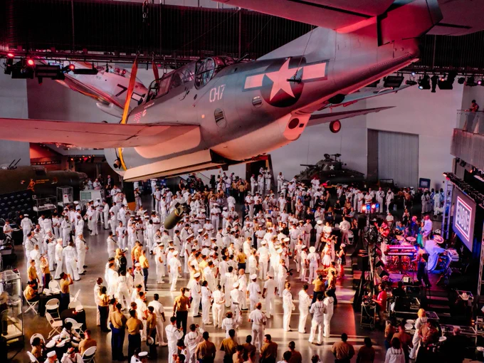 Service members and guests gather inside the National World War II Museum as a live band performs beneath historic aircraft exhibits during a Sail 250 event in New Orleans.