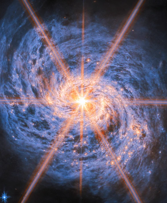 A detailed mid-infrared view of the spiral galaxy Messier 77. The image is dominated by an intensely bright central core, from which six long and two shorter bright rays extend outward like a starburst pattern — these are diffraction spikes caused by the telescope’s optics. Two prominent spiral arms curve outward from the center, marked by glowing orange bubbles of newly formed star clusters. The rest of the galaxy consists of swirling, smoky blue clouds of dust that form intricate filaments and dark cavities throughout the disk.