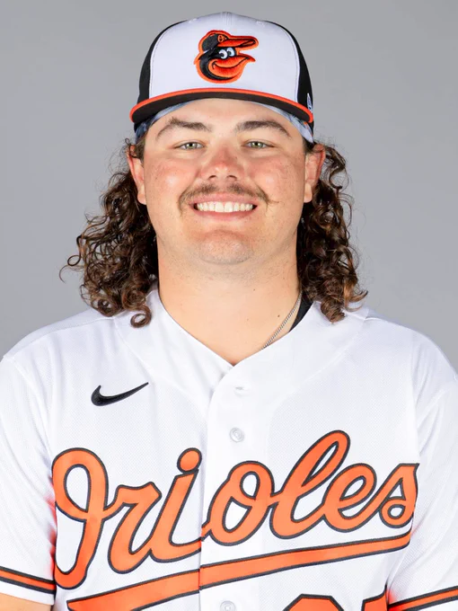 Creed Willems looks at the camera and smiles. He is wearing a white Orioles jersey and a tri-color cap.