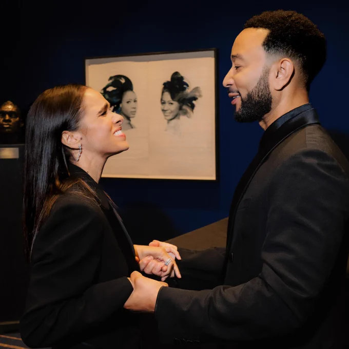 Two people stand facing each other, holding hands and smiling. They are in a room with dark blue walls, featuring a photograph of Josephine Baker in the background.