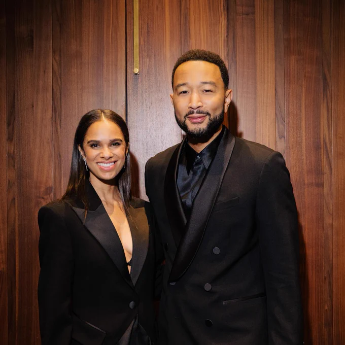 Two people dressed in formal black attire stand together, smiling, in front of a wooden wall.
