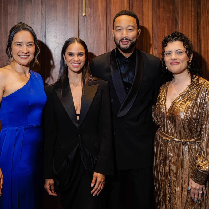 Four people stand together in front of a wooden backdrop, smiling. The person in the blue dress is next to the person wearing a black outfit. Next is the person in the black suit, followed by the person in a shiny gold dress.
