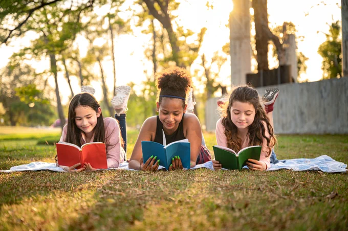 Three young people lay down on the grass in a yard. All three are reading books.