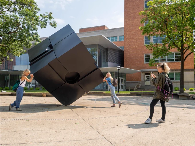 Two people spin a large black cube sculpture while a third person takes a picture