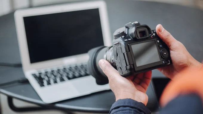 Person holding dslr camera pointing towards opened laptop on table in near background.