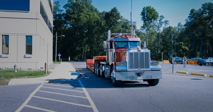 A red Peterbilt truck with a flatbed trailer is parked outside a large beige building on a clear day. The background features trees and several parked cars.