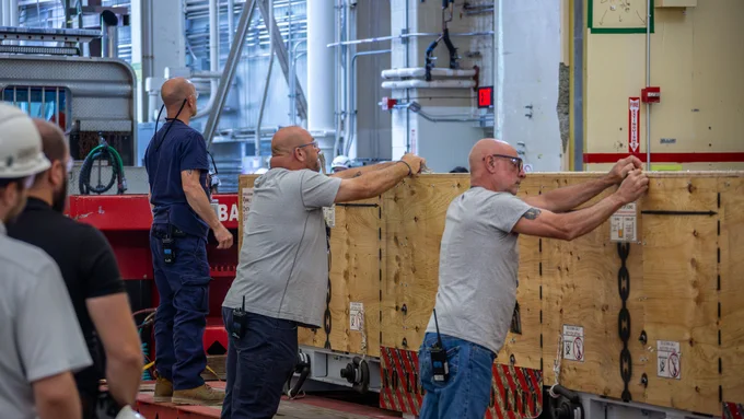 Three people are working together to move a large wooden crate in an industrial setting. They are standing at the crate and marking for unpacking, which has metal reinforcements and appears heavy. In the background, industrial equipment and machinery are visible. The setting is busy and appears to involve teamwork and coordination.