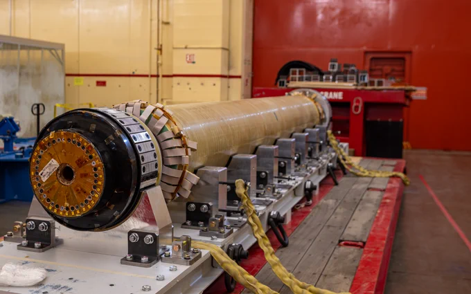 The image shows the central magnet bundle for the National Spherical Torus Experiment-Upgrade resting horizontally on a sturdy frame with clamps securing it in place. The magnet has a cylindrical shape with a complex, detailed end cap and is surrounded by mechanical components. In the background, industrial equipment and machinery are visible in a spacious indoor setting.
