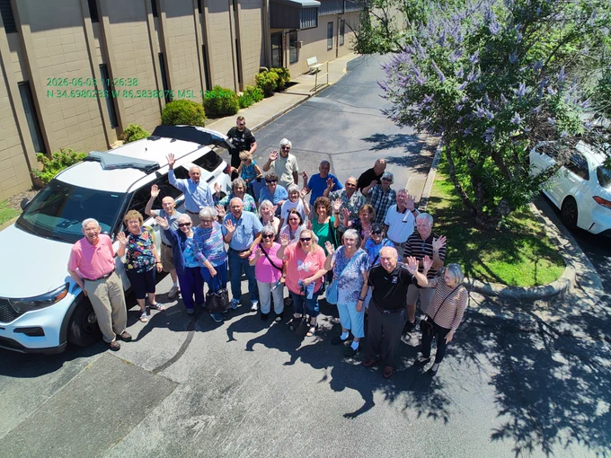 A large group of people is gathered outdoors in a parking lot next to a white SUV. Many individuals are standing close together and waving upward toward the camera. The setting includes a building with tan walls, a paved driveway, a flowering tree casting shadows, and several parked vehicles. Text displaying the date, time, and GPS coordinates appears near the top of the image.
