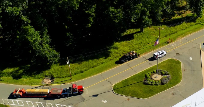 Aerial view of a road with a red semi-truck hauling a long, flatbed trailer. The trailer is carrying large, flat objects. Nearby, a pickup truck and a white SUV are parked. A circular grassy area with an American flag and surrounding plants is visible. Trees line one side of the road.