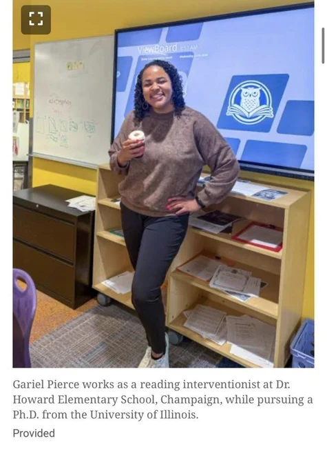 A woman in a brown shirt and black pants holds a donut in a room with shelves.