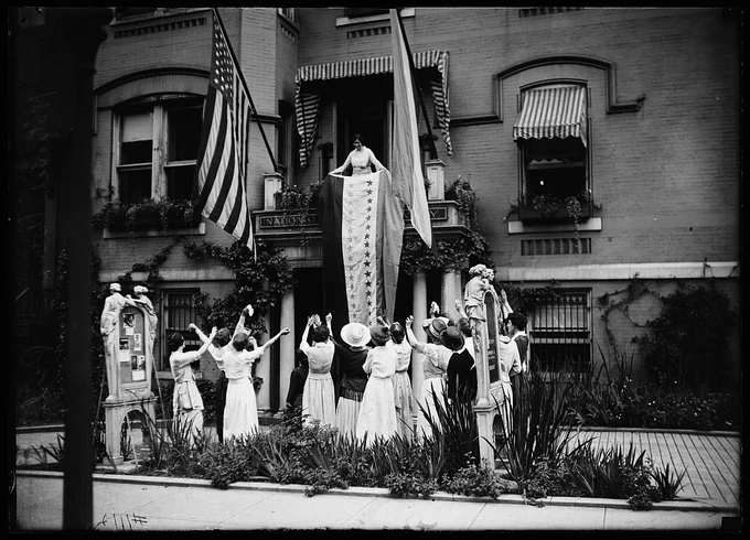 Photo shows a group of women waving their arms in celebration while the ratification banner with its 36 victory stars hangs from the balcony of the National Woman's Party headquarters. 1920. Photo by Harris & Ewing. National Women's Party Records, Library of Congress.