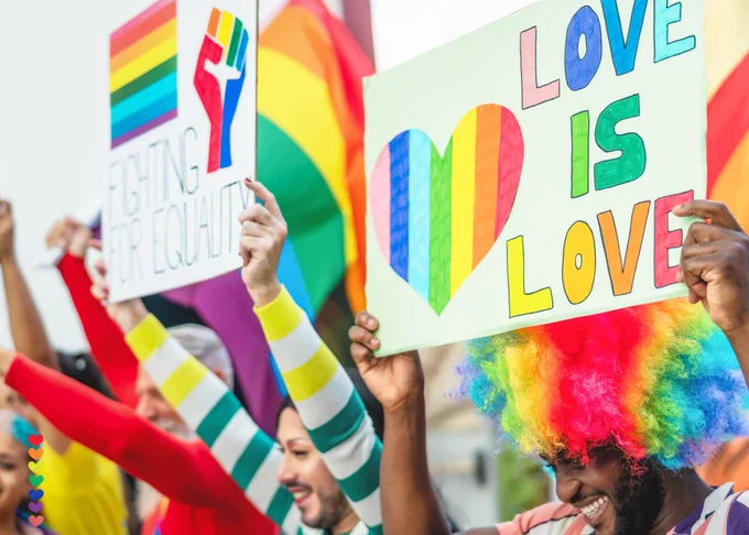 A group of diverse people joyfully holding rainbow-themed signs and flags at a pride event. Signs read "Fighting for Equality" and "Love is Love." One individual wears a rainbow wig.