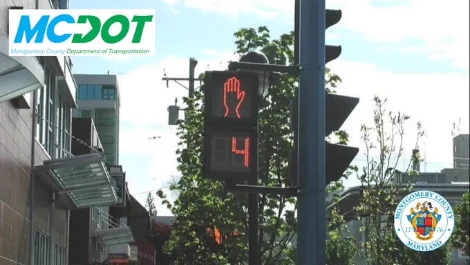 Pedestrian signal showing a red hand and countdown of 4 seconds at a city intersection with buildings and trees in the background. mcdot and county logos