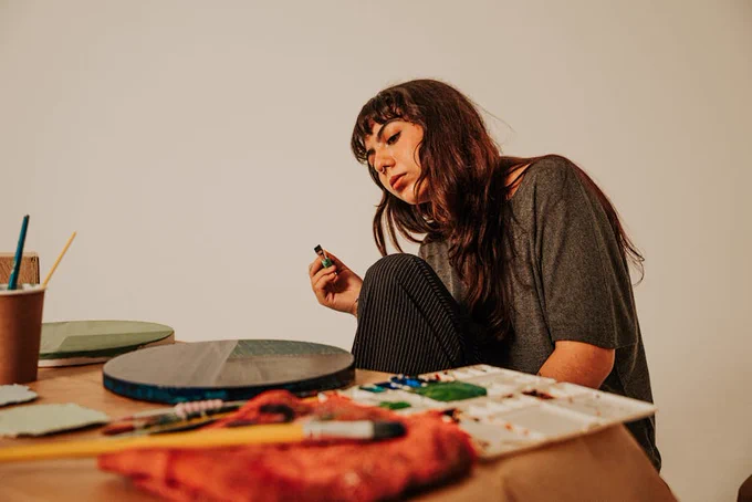 A thoughtful female artist holds a paint bottle while surrounded by her tools in an art studio.