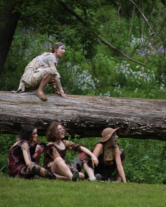 actors in a production of A Midsummer Night’s Dream perch on a tree log and sit in the grass