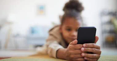 Child lying on the floor holding a smartphone with both hands, focusing on the screen.