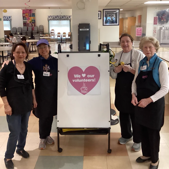 Four volunteers wearing aprons stand around a sign that reads 'We love our volunteers!' inside a community center.