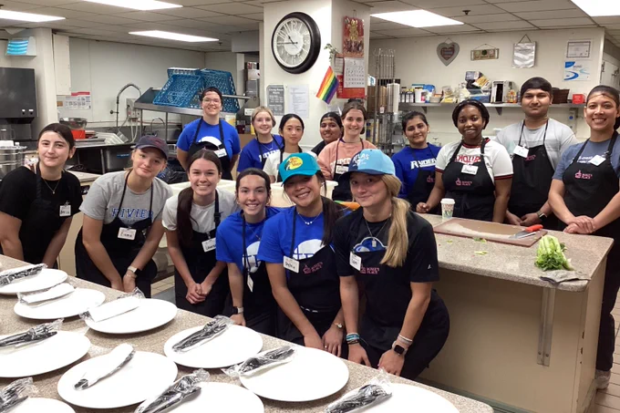 Group of volunteers posing together behind a counter set with plates and utensils in a commercial kitchen.