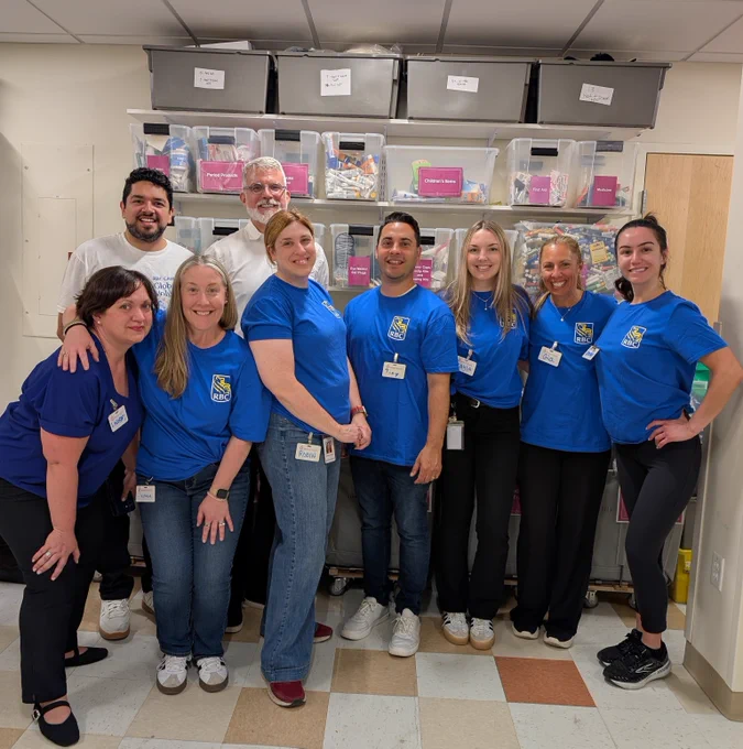 Group of eight people wearing matching blue shirts standing together in a room with organized shelves filled with labeled plastic bins.