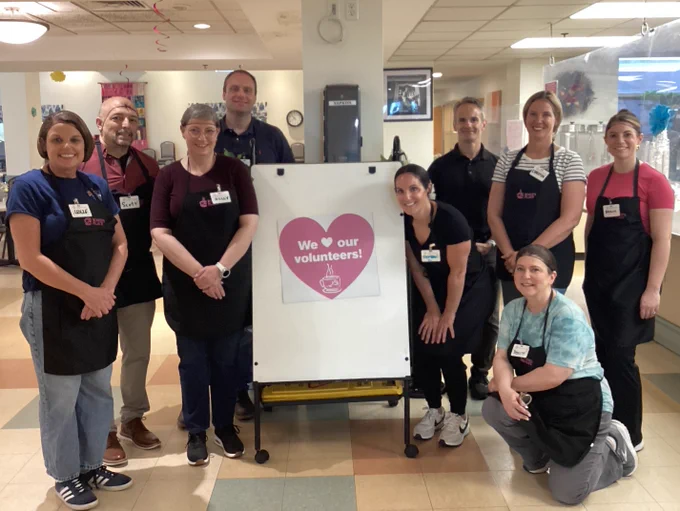 Group of volunteers wearing aprons standing around a sign that says 'We love our volunteers!' inside a community center.
