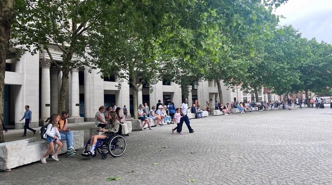 Tree-lined cobblestone plaza beside a stone building, with many people sitting on benches and walking, including a wheelchair user.