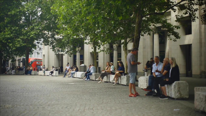People sit on stone benches beneath leafy trees along a cobblestone plaza beside a columned building; a red double-decker bus passes behind.