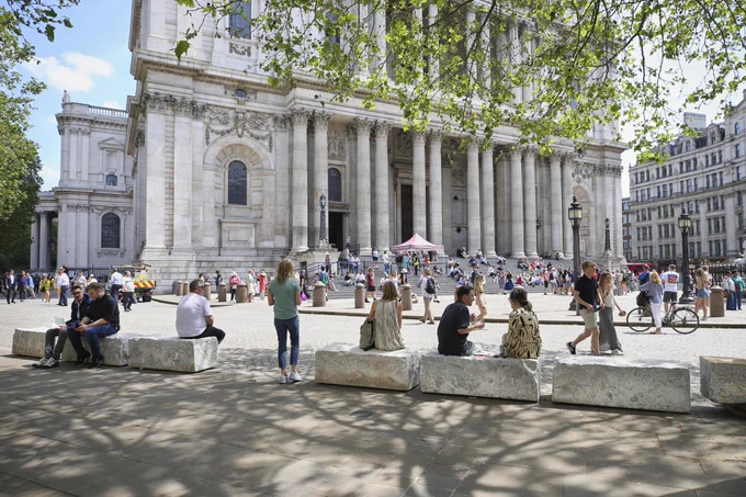 People relax and gather outside St. Paul’s Cathedral in London, beneath leafy trees on a bright day.