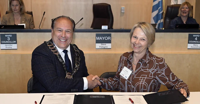 Two individuals, from left: Mayor Steve Pelligrini, King Township, and President and Vice-Chancellor Lisa Philipps, York University, sign the MOU at a table, shaking hands in a formal setting. Behind them, others are seated, some smiling. Documents are placed on the table in front of them.