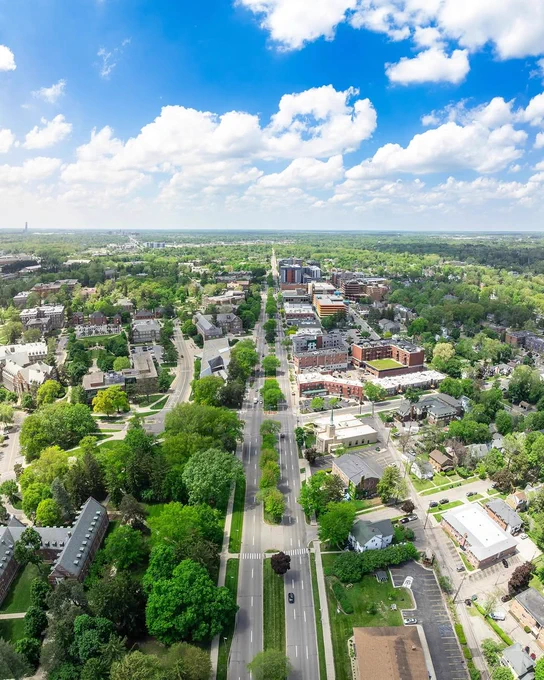 Aerial view of East Lansing and Michigan State University on a sunny day, showing tree-lined streets, campus buildings, and a blue sky filled with scattered white clouds.
