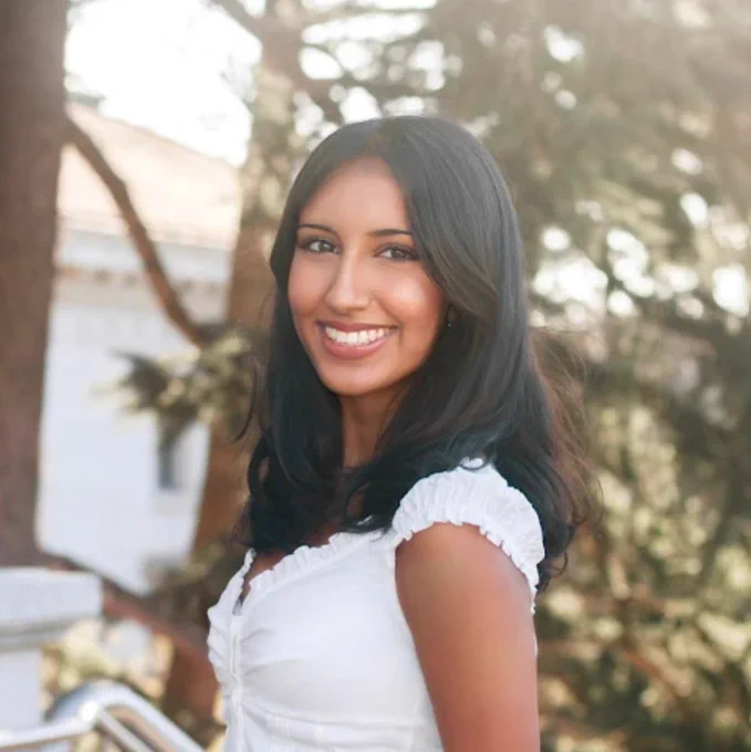 A woman with long dark hair smiles warmly in an outdoor setting. She wears a white top, with soft sunlight filtering through trees in the background.