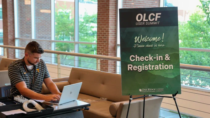Staff member working on a laptop beside a sign welcoming participants to the Oak Ridge Leadership Computing Facility User Summit registration area inside Oak Ridge National Laboratory.