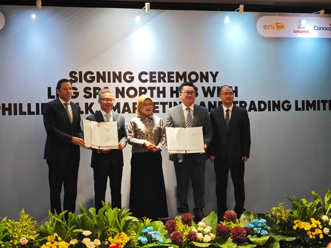 Five individuals are standing on a stage at a signing ceremony, holding documents. The backdrop displays logos of Eni, Sinopec, and ConocoPhillips, with a text saying "Signing Ceremony LHS SR3 North Hibiscus with Phillips Black Marketing And Trading Limited." There are plants arranged at the front of the stage.
