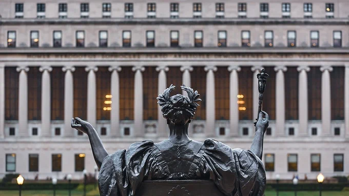 The image shows a statue positioned in front of the iconic facade of Butler Library at Columbia University. The statue is facing the building, holding objects in both hands. The library's grand columns and large windows are visible in the background.