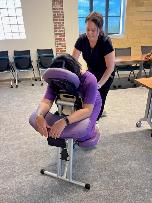 A person is seated in a massage chair receiving a massage from another person in an office setting. The massage chair is purple. There are tables and chairs in the background.