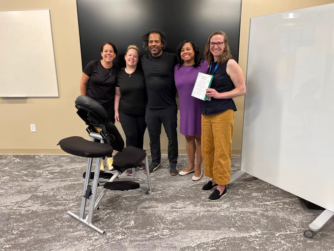 A group of five people standing together in a room with a massage chair in the foreground. One person holds a document. There are whiteboards and a large black screen in the background. The setting appears to be an office or professional environment.