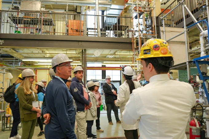 A person in a lab coat and hard hat speaks to a group wearing hard hats in a multi-level laboratory filled with pipes and equipment.