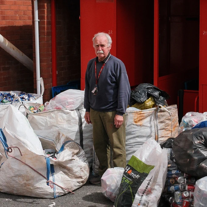 An older man wearing a dark sweatshirt and lanyard stands among large bags of recyclables and trash outside a brick building with bright red doors. Crushed aluminum cans and plastic bags are piled around him in an industrial waste-sorting area.