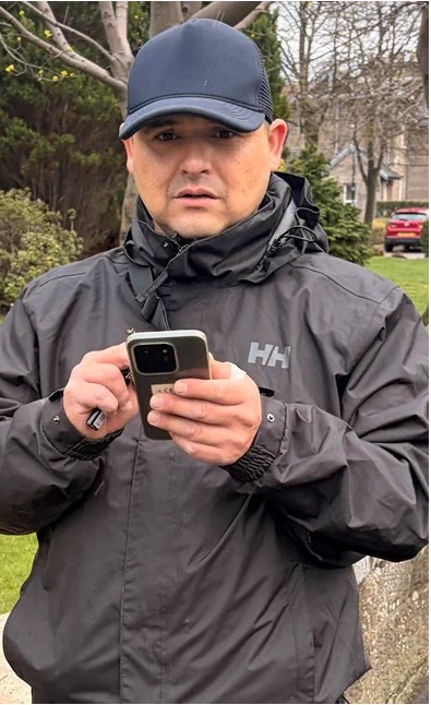 Man holding a mobile phone in front of him. He is wearing a black baseball cap and black waterproof jacket. Behind him are trees, houses and a red car.