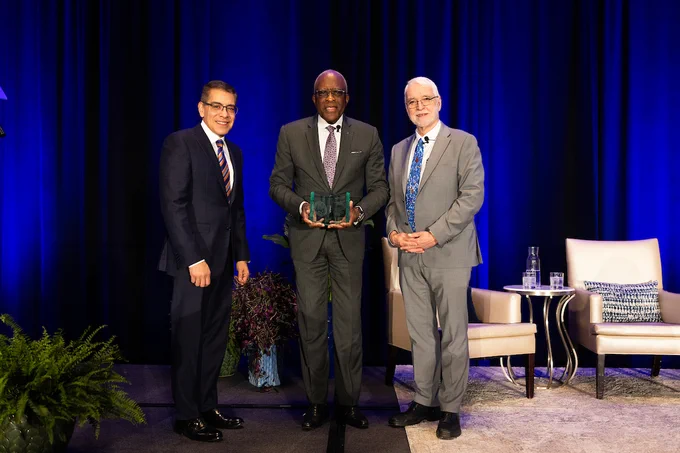Three men in suits stand on a stage with dark blue curtains, the center man holding a glass award between two seated chairs and a small table.