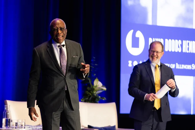 Two men in suits speaking on stage during a formal event with a blue backdrop and a screen displaying text.