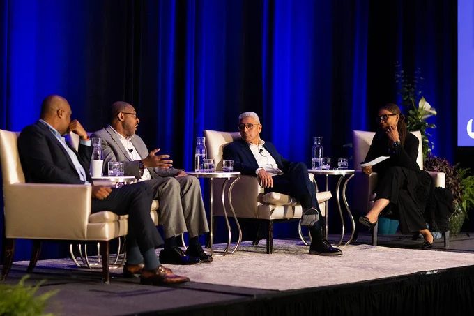 Four professionals seated on stage engaged in a panel discussion with blue curtains and water glasses on side tables.