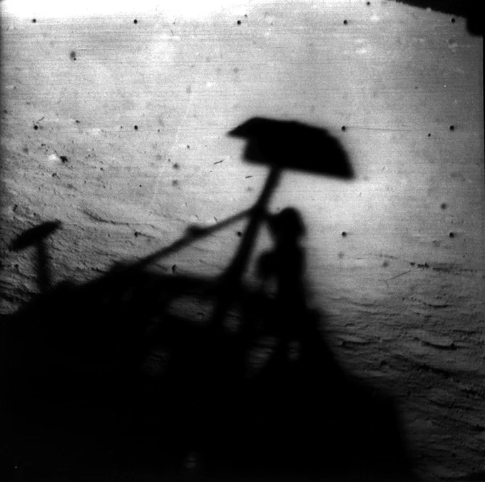 Black and white image of the surface of the Moon showing the shadow of the Surveyor I spacecraft that took it in the late lunar afternoon. The horizon is at the upper right.