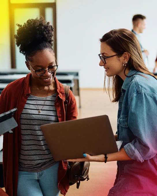 Alt text: Two students stand together in a bright campus space, smiling as they review information on a laptop. One student holds the computer while the other looks on, creating a collaborative and welcoming learning environment.