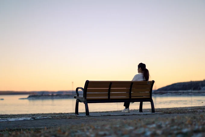 A person sits alone on a wooden bench facing a calm waterfront at sunset, with a soft yellow sky and distant shoreline in the background.