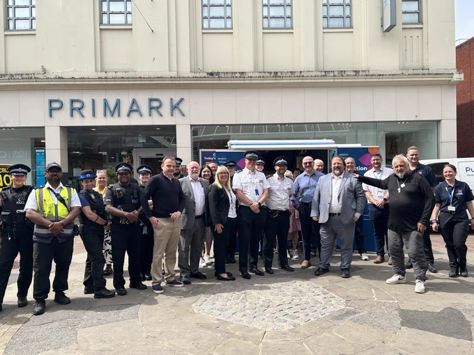 Police officers and civilians stand together outside a store, with a branded van in the background.