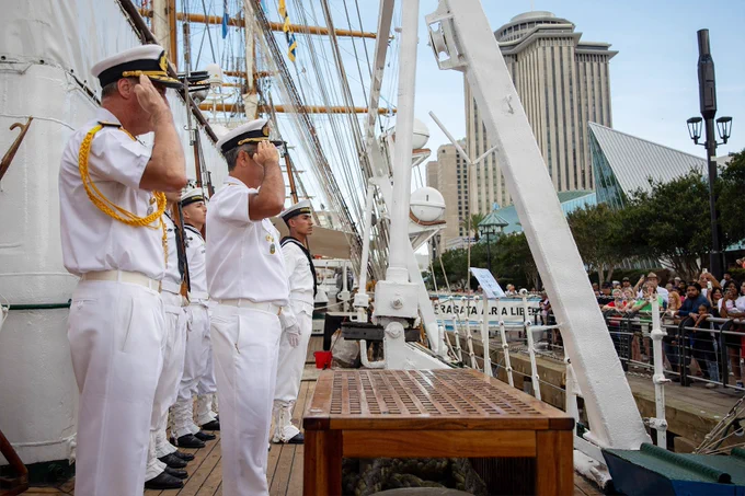 Argentine naval personnel render honors from the deck of a tall ship while visitors gather along the waterfront during a public maritime event.